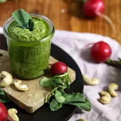 Frisches Radieschen Pesto in einem Glas, umgeben von Radieschen, Cashewkernen und grünen Blättern auf einem Holztisch.
