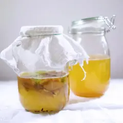 Zwei Gläser mit gelblicher Flüssigkeit. Im vorderen Glas schwimmen Apfelstücke unter einem weißen Tuch. Hier wird Apfelessig selber gemacht.