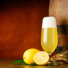 Ein Glas Radler steht neben Zitronen und Gerstenähren vor einem Holzfass.