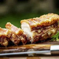 Ein perfekt gebratener, knuspriger Schweinebauch mit goldbrauner Kruste, auf einem Holzbrett mit Petersilie und einem Messer.