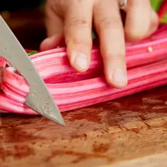 Eine Hand schneidet mit einem Messer die roten mangoldstiele auf einem Holzbrett zu.