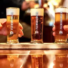 Drei Gläser Flensburger Bier mit cremigem **Bier schaum** stehen auf einem Holztablett. Hände greifen nach den Gläsern.