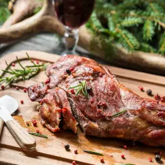 Ein saftiger Rehschlegel, perfekt gegrillt, liegt auf einem Holzbrett. Gewürze und Rosmarin zieren das Fleisch. Ein Glas Rotwein und Hirschgeweih im Hintergrund deuten auf ein festliches rehschlegel rezept hin.