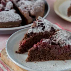 Saftiger Schoko-Kirsch-Kuchen aus der Springform, mit Puderzucker bestäubt. Zwei Stücke auf einem Teller, der ganze Kuchen im Hintergrund.