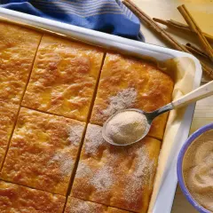 Goldbrauner Kartoffelkuchen vom Blech, frisch aus dem Ofen, wird mit Zimt-Zucker bestreut. Daneben liegen Zimtstangen und eine Schüssel mit der süßen Mischung.