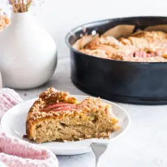 Ein Stück Apfelkuchen mit Haselnuss-Streuseln auf einem Teller, daneben eine Backform mit dem restlichen Kuchen.
