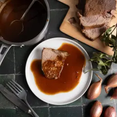 Ein Stück Rinderbraten mit aromatischer Rinderbraten Sauce auf einem Teller, daneben ein Topf mit Sauce, ein Messer und eine Gabel.