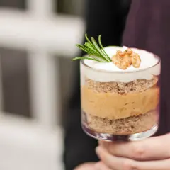 Ein Glas mit Schichten von Kuchen und cremiger Karamell-Mousse, garniert mit Rosmarin und einer Walnusshälfte.