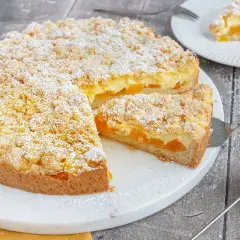 Saftiger aprikosenkuchen mit pudding und Streuseln, bestäubt mit Puderzucker. Ein Stück wird gerade serviert.