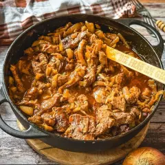 Saftiger Zwiebelbraten in einer Gusseisenpfanne mit Zwiebeln und Fleisch, daneben halbierte Zwiebeln.