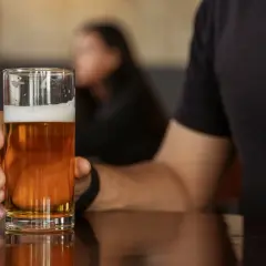 Mann hält ein Glas mit hellem englischem Bier. Der Schaum krönt das goldene Getränk, während im Hintergrund eine Person verschwommen zu sehen ist.