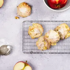 Saftige Apfelkekse, frisch gebacken und mit Puderzucker bestäubt, liegen auf einem Kuchengitter. Äpfel und Apfelschale sind daneben arrangiert.