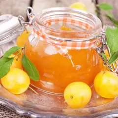 Ein Glas mit leuchtend orangefarbener Mirabellenmarmelade, umgeben von frischen Früchten und Blättern. Ein schönes Bild für ein Mirabellenmarmelade Rezept.