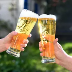 Zwei Hände halten Gläser mit Bier für einen Bier Vergleich. Das goldene Bier mit Schaumkrone und Weizenähren-Dekor glänzt in der Sonne.