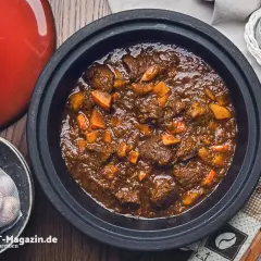 Herzhaftes Rehhals Rezept in einem Schmortopf, mit Karottenstücken und Knoblauch daneben.
