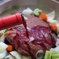 Rinderbraten in Rotwein einlegen. Ein roter Weinstrahl ergießt sich über das Fleisch, umgeben von Karotten, Sellerie und Zwiebeln.