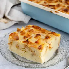 Ein Stück von Omas Brotpudding, goldbraun gebacken, auf einem Glas-Teller. Im Hintergrund ein Auflaufform und ein Brötchen.