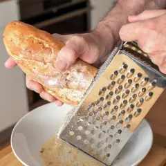 Mann reibt ein Baguette über einer Schüssel, um Paniermehl selber zu machen. Frische Brösel für die Küche.