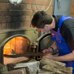 Mann legt Holz in den Ofen für die Rauchbier Herstellung. Das Feuer glüht hell.
