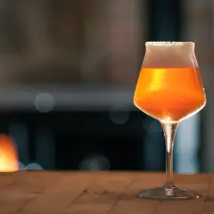 Ein Glas mit bernsteinfarbenem Bier, das nach pale ale malz duftet, steht auf einem Holztisch. Im Hintergrund glüht ein Kaminfeuer.