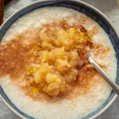 Cremiger Milchreis mit Apfelmus und Zimt, der weltbester Milchreis-Geschmack verspricht. Daneben liegen Zimtstangen.