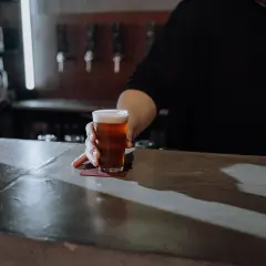 Ein Glas Bier wird über den Tresen geschoben. Der Unterschied zwischen Bier und Pils ist hier nicht ersichtlich, aber die Farbe des Bieres ist bernsteinfarben.