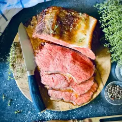 Saftiger Rostbraten, perfekt im Ofen zubereitet, liegt auf einem Holzbrett. Daneben liegen Kräuter, Salz und Pfefferkörner.