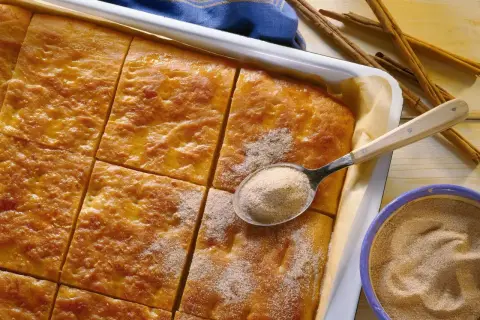 Goldbrauner Kartoffelkuchen vom Blech, frisch aus dem Ofen, wird mit Zimt-Zucker bestreut. Daneben liegen Zimtstangen und eine Schüssel mit der süßen Mischung.