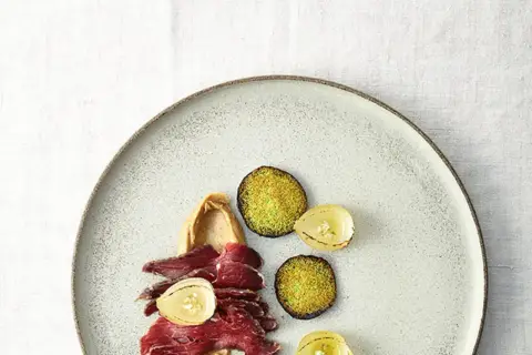 Gebeiztes Rinderfilet mit cremiger Beilage, karamellisierten Zwiebeln und grünen, knusprigen Scheiben auf einem Teller.