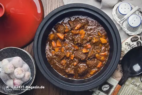 Herzhaftes Rehhals Rezept in einem Schmortopf, mit Karottenstücken und Knoblauch daneben.