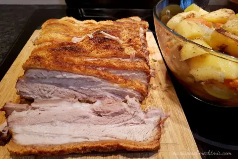 Ein butterzarter Schweinebauch mit knuspriger Kruste liegt auf einem Holzbrett, daneben eine Schüssel mit Bratkartoffeln und Gemüse.