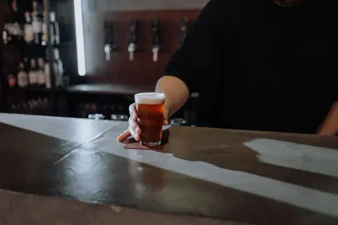 Ein Glas Bier wird über den Tresen geschoben. Der Unterschied zwischen Bier und Pils ist hier nicht ersichtlich, aber die Farbe des Bieres ist bernsteinfarben.