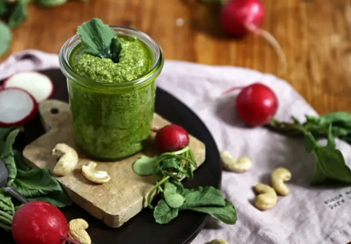 Frisches Radieschen Pesto in einem Glas, umgeben von Radieschen, Cashewkernen und grünen Blättern auf einem Holztisch.