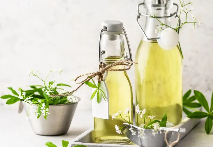 Zwei Flaschen mit omas waldmeistersirup, verziert mit frischen Waldmeisterzweigen und kleinen weißen Blüten.