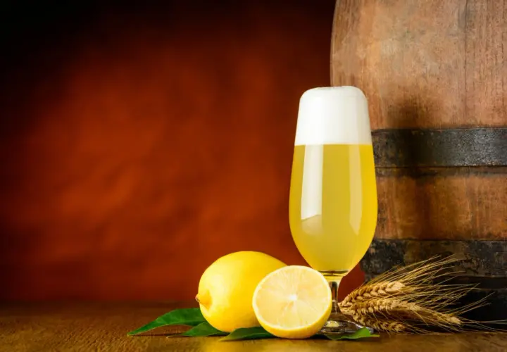 Ein Glas Radler steht neben Zitronen und Gerstenähren vor einem Holzfass.