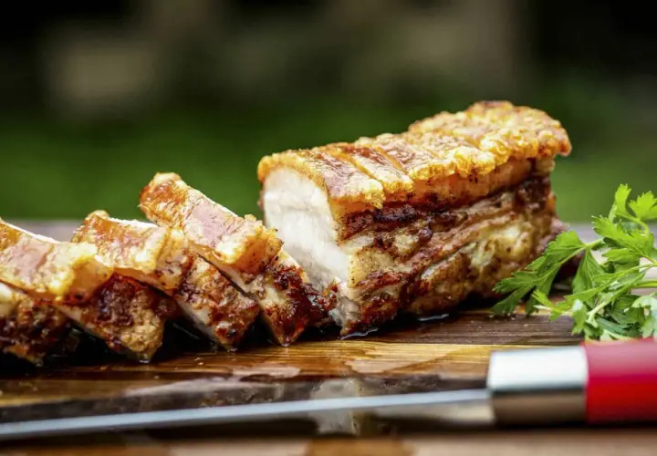 Ein perfekt gebratener, knuspriger Schweinebauch mit goldbrauner Kruste, auf einem Holzbrett mit Petersilie und einem Messer.