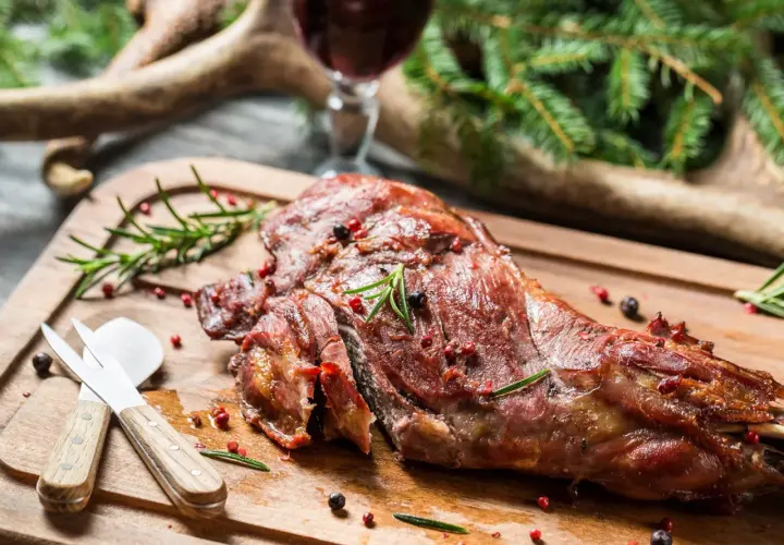Ein saftiger Rehschlegel, perfekt gegrillt, liegt auf einem Holzbrett. Gewürze und Rosmarin zieren das Fleisch. Ein Glas Rotwein und Hirschgeweih im Hintergrund deuten auf ein festliches rehschlegel rezept hin.