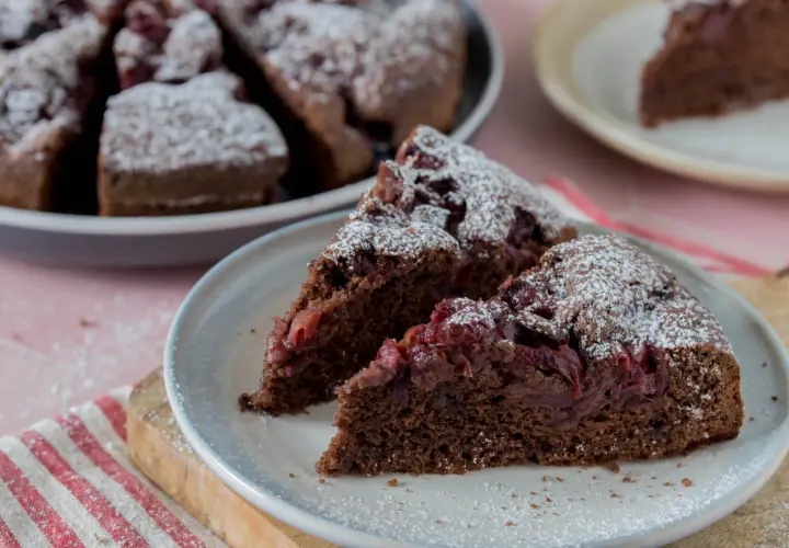 Saftiger Schoko-Kirsch-Kuchen aus der Springform, mit Puderzucker bestäubt. Zwei Stücke auf einem Teller, der ganze Kuchen im Hintergrund.