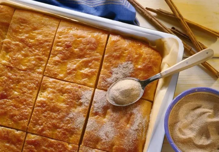 Goldbrauner Kartoffelkuchen vom Blech, frisch aus dem Ofen, wird mit Zimt-Zucker bestreut. Daneben liegen Zimtstangen und eine Schüssel mit der süßen Mischung.