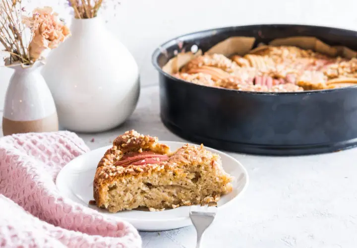 Ein Stück Apfelkuchen mit Haselnuss-Streuseln auf einem Teller, daneben eine Backform mit dem restlichen Kuchen.