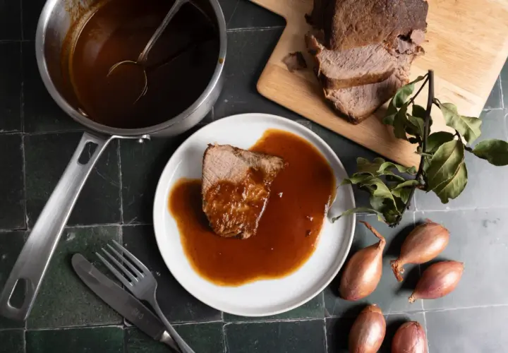 Ein Stück Rinderbraten mit aromatischer Rinderbraten Sauce auf einem Teller, daneben ein Topf mit Sauce, ein Messer und eine Gabel.