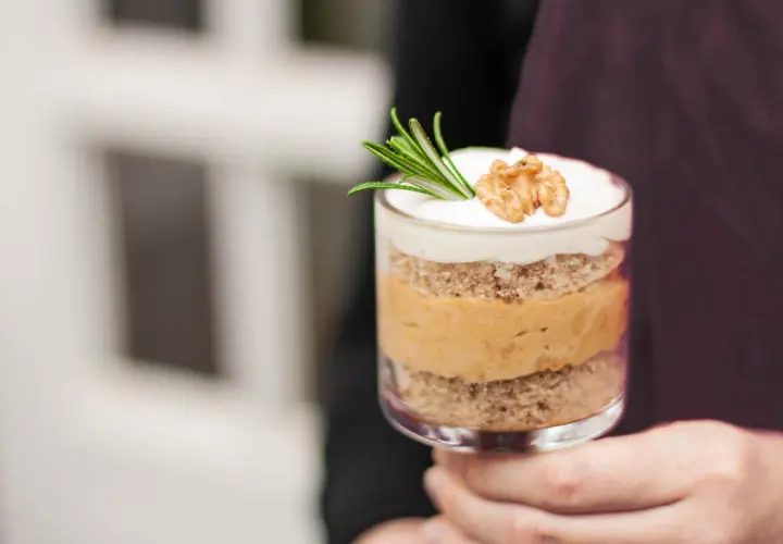 Ein Glas mit Schichten von Kuchen und cremiger Karamell-Mousse, garniert mit Rosmarin und einer Walnusshälfte.
