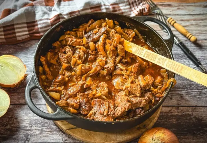 Saftiger Zwiebelbraten in einer Gusseisenpfanne mit Zwiebeln und Fleisch, daneben halbierte Zwiebeln.