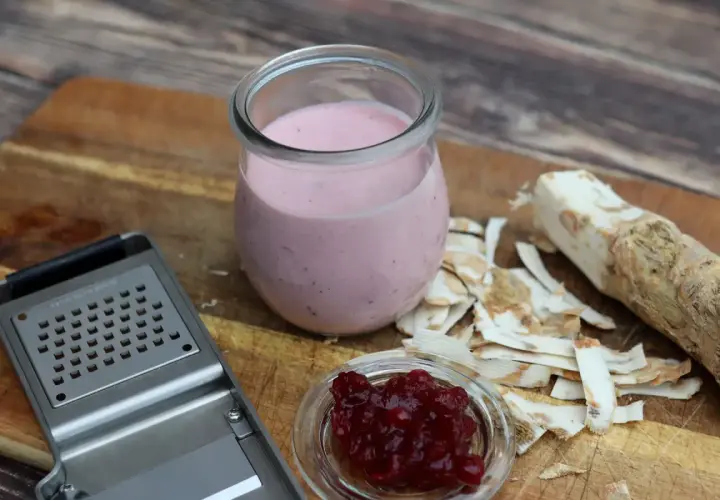 Meerrettichschmand in einem Glas, daneben frisch geriebener Meerrettich, eine Reibe und Preiselbeeren.