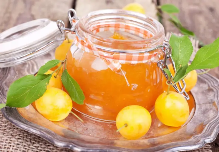 Ein Glas mit leuchtend orangefarbener Mirabellenmarmelade, umgeben von frischen Früchten und Blättern. Ein schönes Bild für ein Mirabellenmarmelade Rezept.