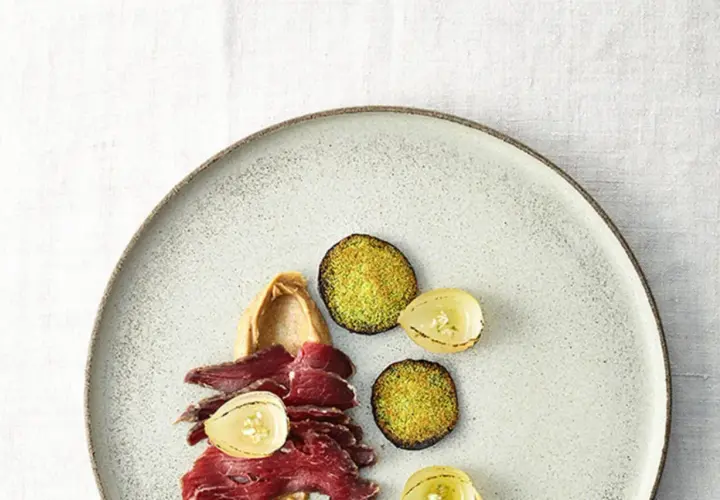 Gebeiztes Rinderfilet mit cremiger Beilage, karamellisierten Zwiebeln und grünen, knusprigen Scheiben auf einem Teller.