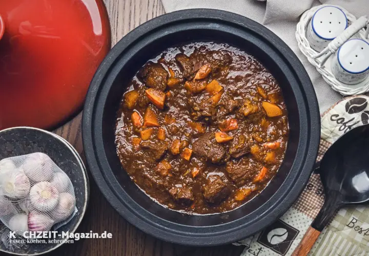 Herzhaftes Rehhals Rezept in einem Schmortopf, mit Karottenstücken und Knoblauch daneben.