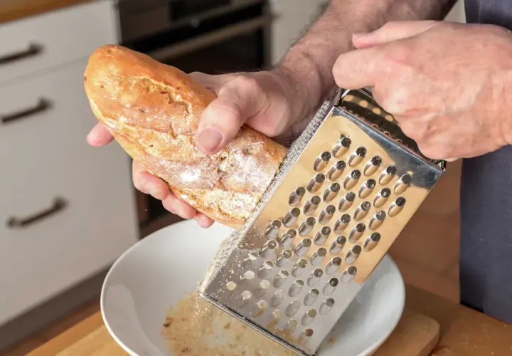 Mann reibt ein Baguette über einer Schüssel, um Paniermehl selber zu machen. Frische Brösel für die Küche.