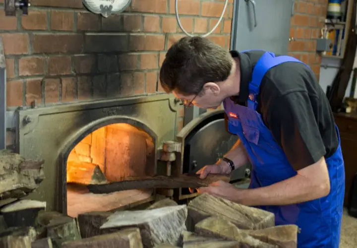 Mann legt Holz in den Ofen für die Rauchbier Herstellung. Das Feuer glüht hell.