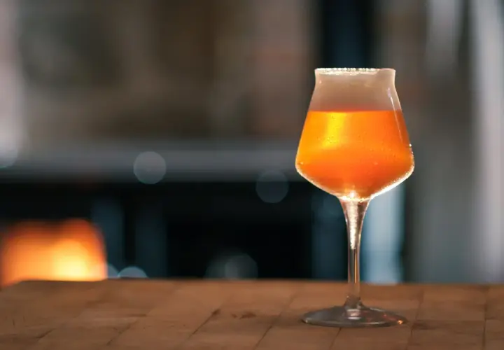 Ein Glas mit bernsteinfarbenem Bier, das nach pale ale malz duftet, steht auf einem Holztisch. Im Hintergrund glüht ein Kaminfeuer.